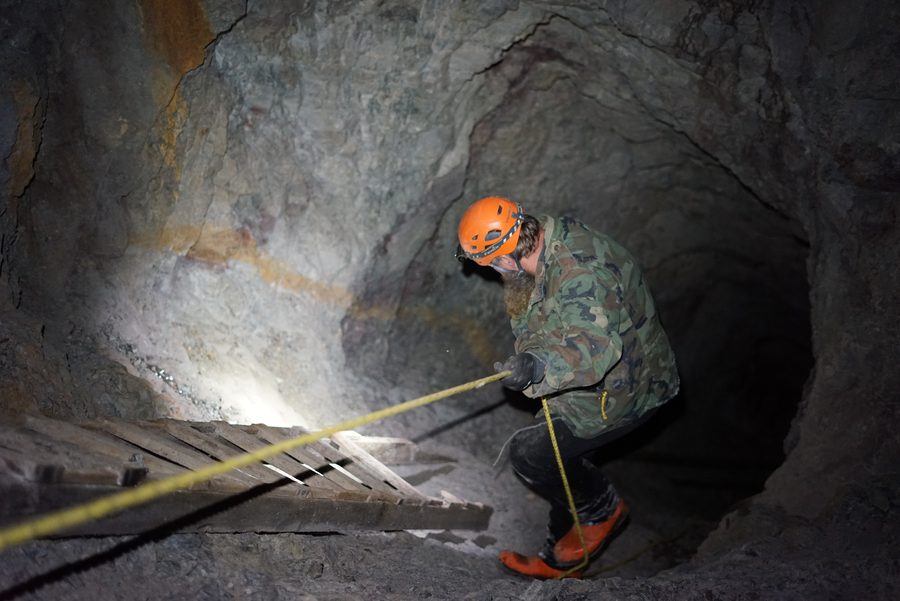 Frank in a camouflage jacket and orange helmet going down a steep rock passage on a yellow rope, beside an old wooden ladder lying on the slope