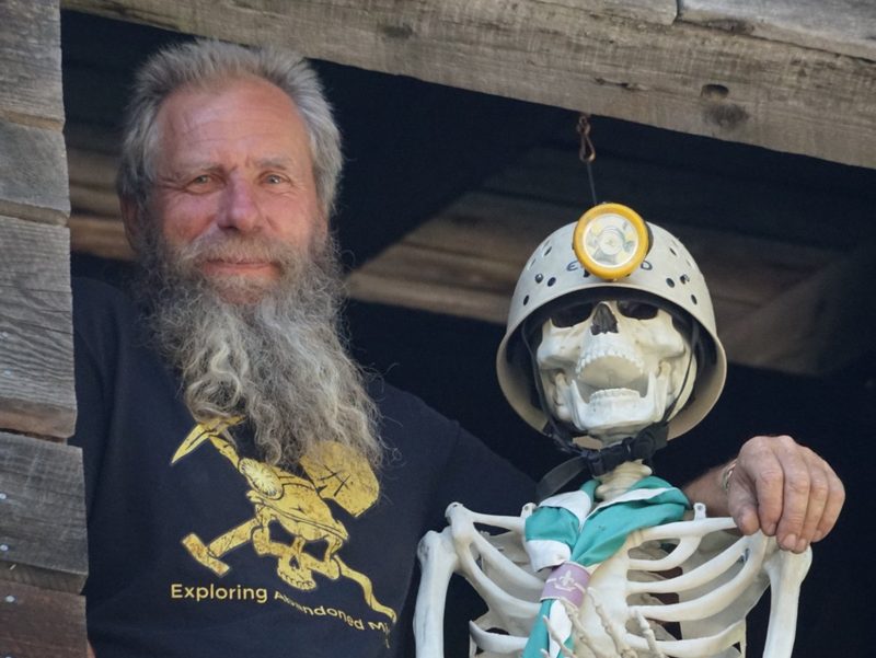 Frank Schlichting smiling from the window of an old wooden building, beside a skeleton in a miner&rsquo;s helmet and lamp