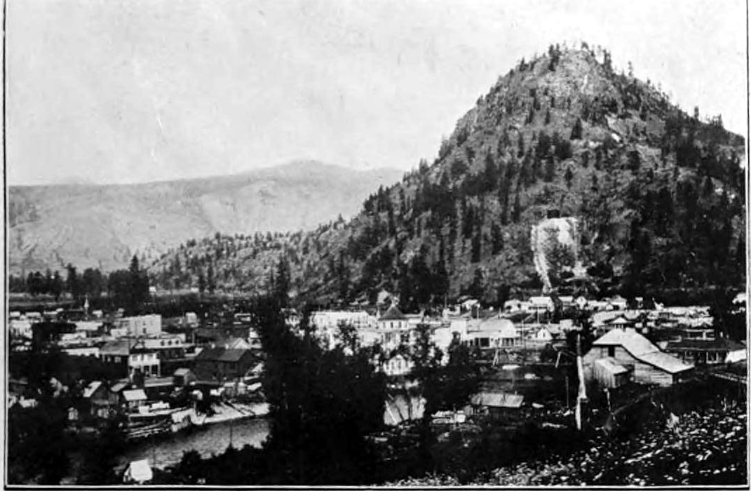 A halftone photograph of Grand Forks, BC, in 1900: frame buildings along the river under a steep forested hill, with bare mountains behind