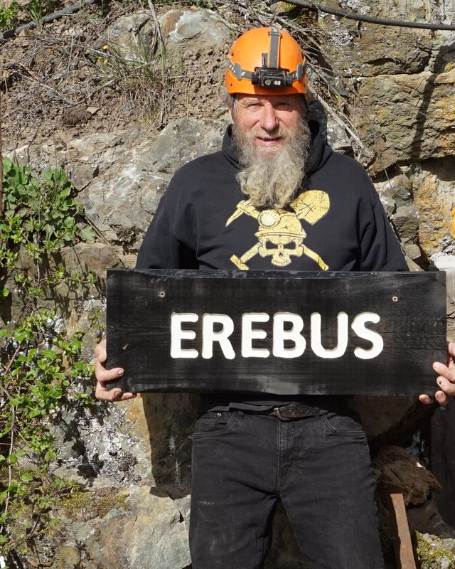 Frank Schlichting in a miner's helmet at the portal, holding up a black sign that reads Erebus
