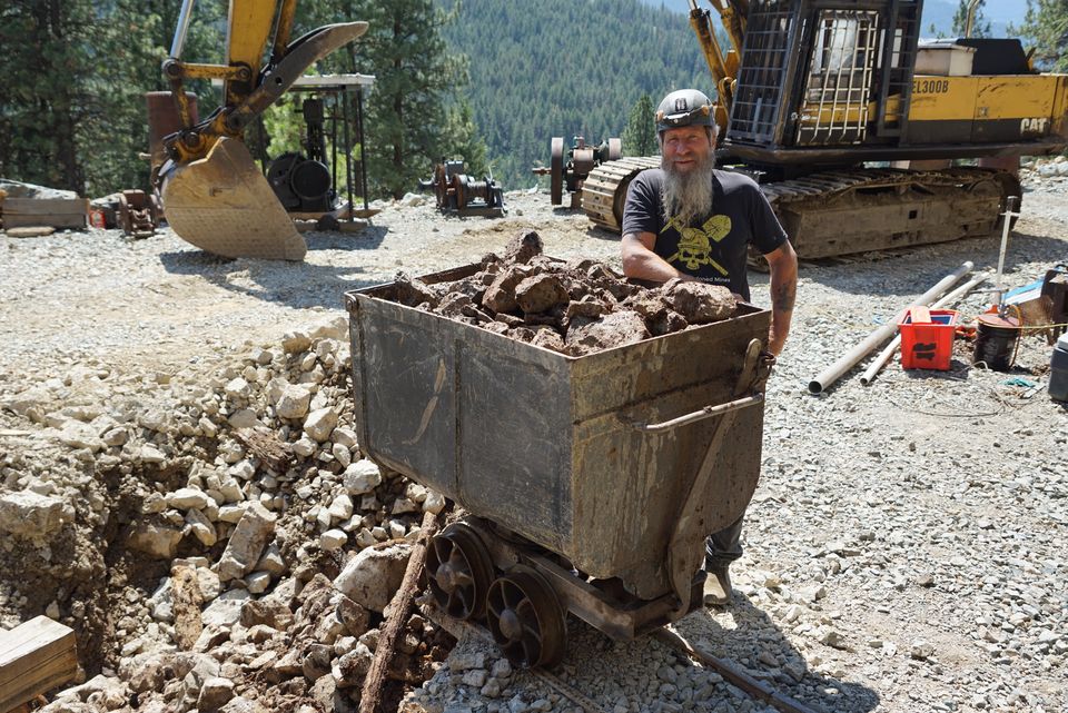 Frank Schlichting outside the mine beside an ore car heaped with rock, an excavator and old mining machinery behind him and forested mountains beyond