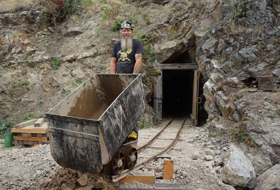 Frank Schlichting, smiling, behind an empty ore car on the rails outside the timbered portal of the Yankee Boy mine