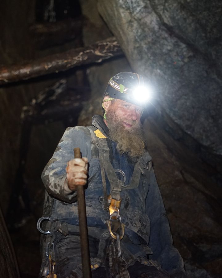 Frank Schlichting underground in a climbing harness, headlamp blazing, gripping a steel bar